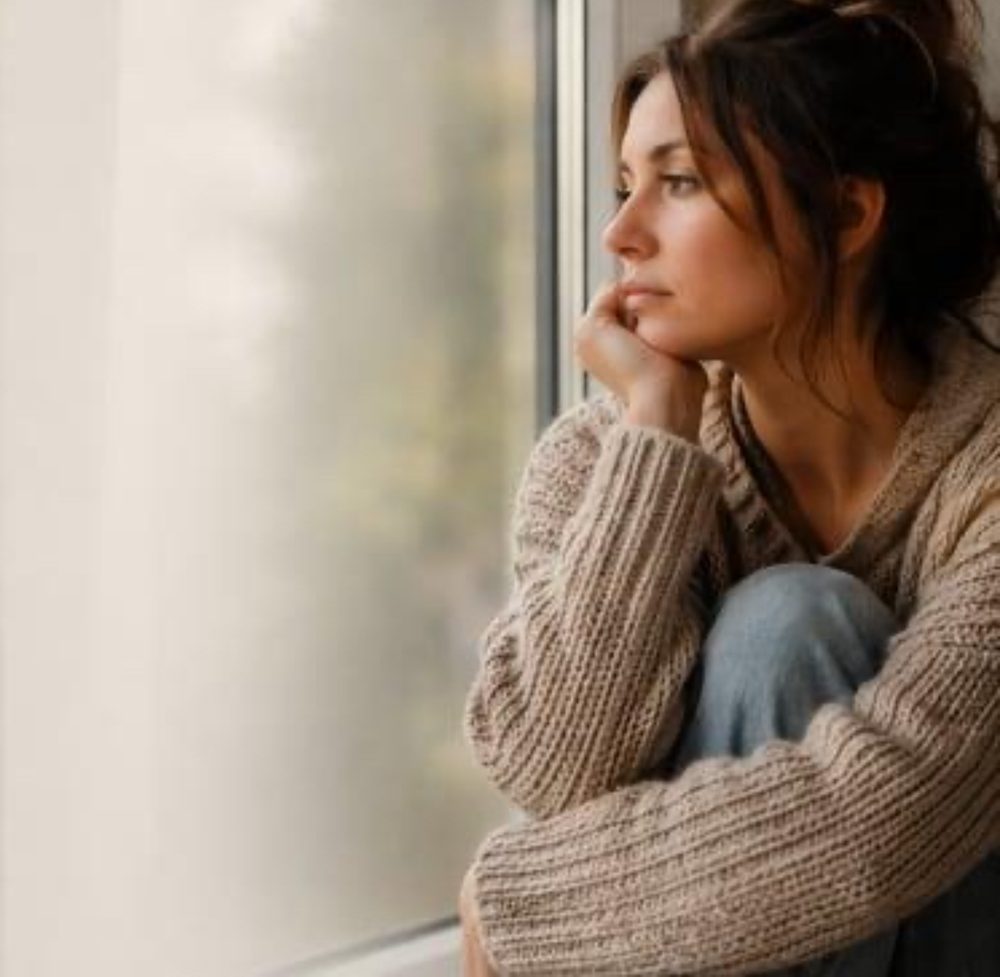 Femme assise près d’une fenêtre, pensive, dans une ambiance calme et naturelle